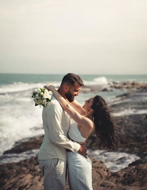 Casal abraçado nas pedras à beira-mar, com buquê.